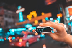 A hand holds up a particulate matter air sensor in front of a blurred background of city traffic