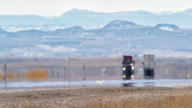 A hot road in an arid environment with visible heat mirage effect. There are two tractor trailers visible in the distance.