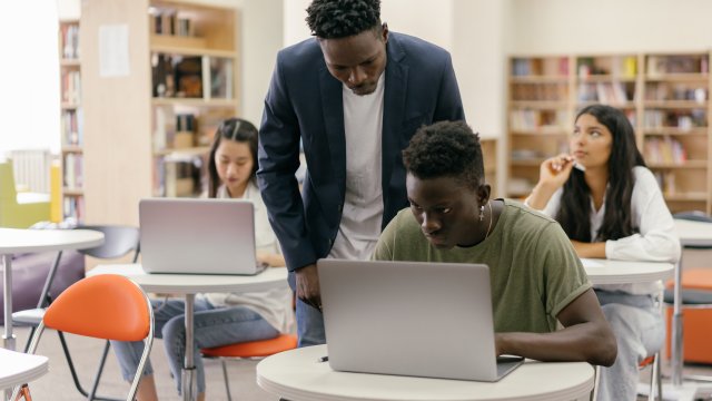 Teacher and three students in a classroom with laptops open in front of them.