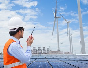 Man in personal protective equipment with a radio looking toward a large, industrial sized windmill.