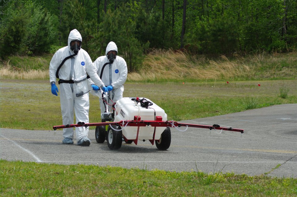 Two team members wearing tyvek suits walk down concrete road using a boom sprayer to inoculate the area.