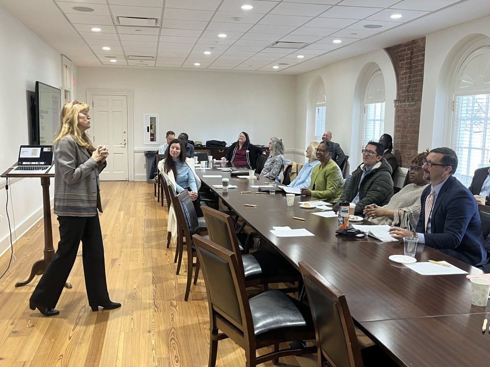 A woman is seen is presenting in front of the room full of people at a table.