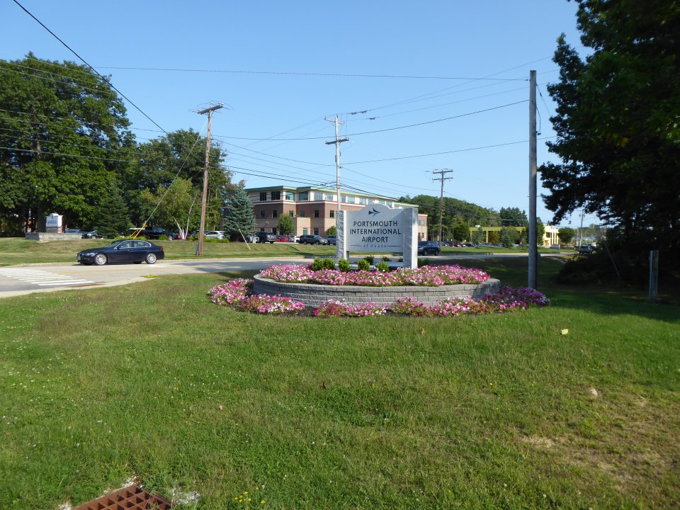 A sign for the Portsmouth International Airport surrounded by flowers and grass, next to a road.