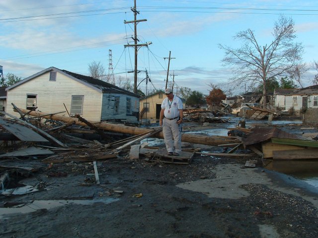 A man stands in the middle of a neighborhood street that is covered in wood and debris from a storm.