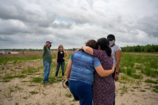 Two women facing away from the camera console each other as they look on a vacant piece of land. Three other people are seen standing around and conversing.