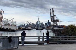 People standing on a low bridge on Lower Spokane Street over the river as it opens up into the estuary with downtown Seattle in the distance.