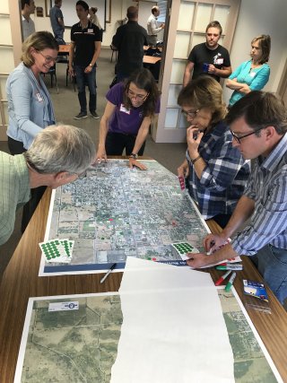 A group of people are standing around a table with a map on it, during a community workshop. 