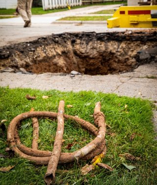 Large hole in sidewalk with a coiled up lead service line that has been removed laying the in grass in front of it.