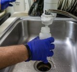 Gloved hand collecting a kitchen tap water sample in a bottle.