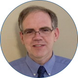 Speaker Matthew Magnuson has dark hair and is wearing eyeglasses with a blue button down shirt while standing against a white background.