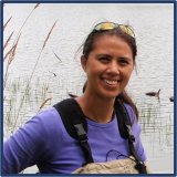 A woman with brown hair in a ponytail. She is wearing waders and is standing at the edge of a lake.