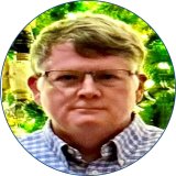 Joseph Wood has light hair and is wearing glasses with a blue checkered collared shirt standing in front of greenery.